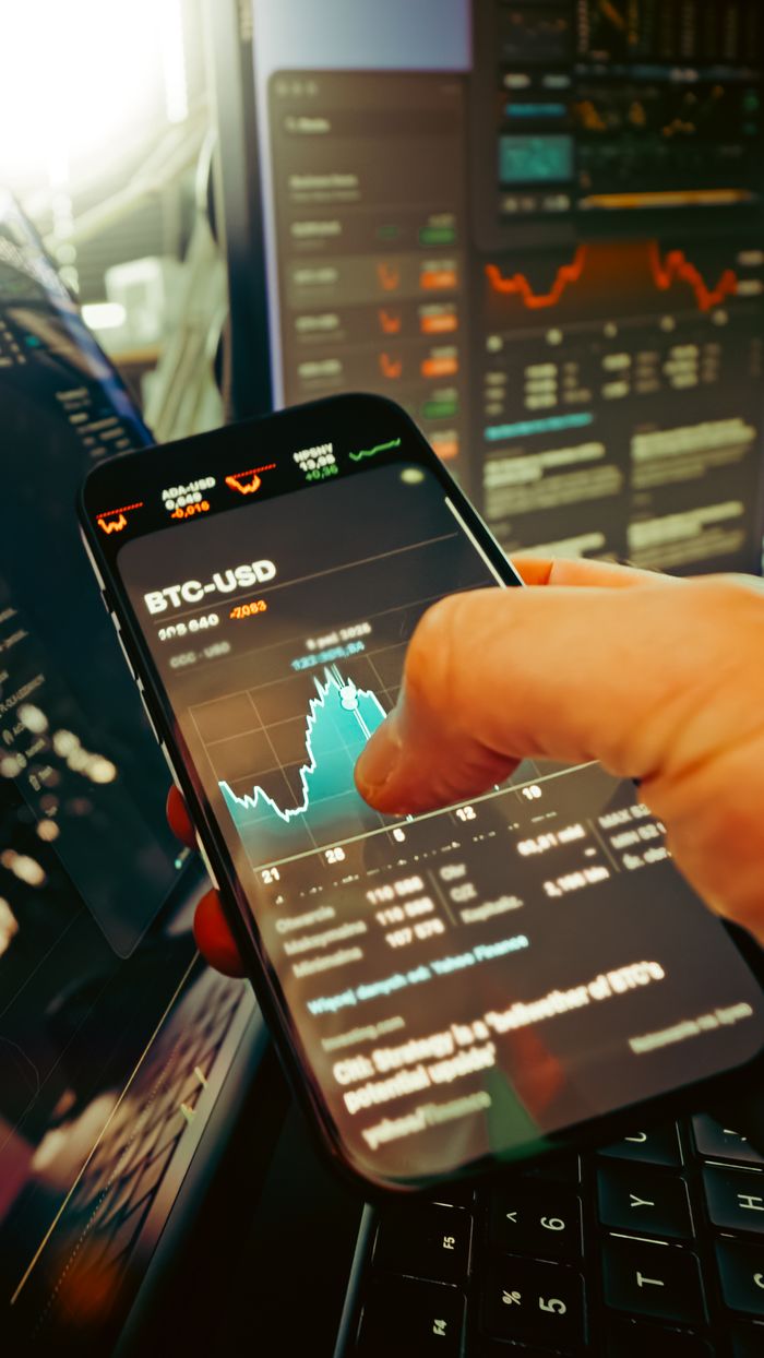 cta-bg A hand holds a smartphone displaying BTC-USD chart amidst digital trading screens, highlighting crypto finance.